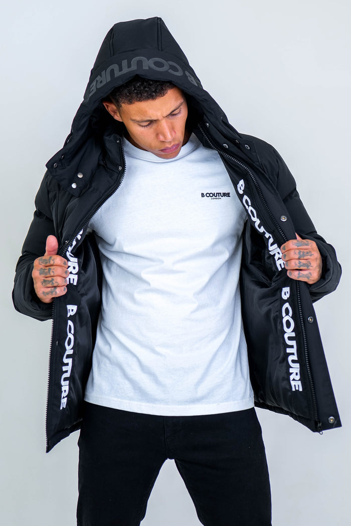 A man with short hair and tattoos on his hands holds open an Arctic Puffer Jacket - Black, revealing its B COUTURE inner print over a white B COUTURE t-shirt—a bold winter outerwear choice.