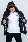 A man with short hair and tattoos on his hands holds open an Arctic Puffer Jacket - Black, revealing its B COUTURE inner print over a white B COUTURE t-shirt—a bold winter outerwear choice.