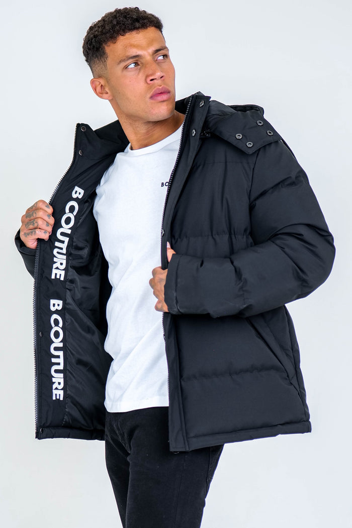 A man in an Arctic Puffer Jacket - Black stands against a plain light background, holding his jacket open to reveal the B COUTURE text inside, showcasing its thermal insulation and winter-ready design.