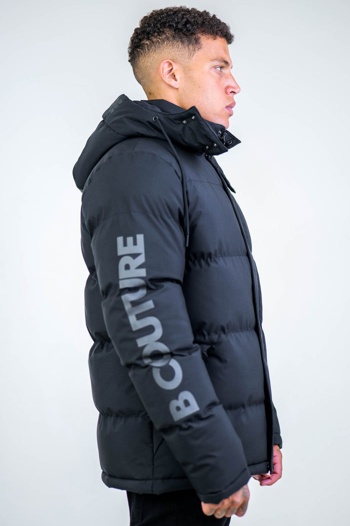 A man in profile wears the Arctic Puffer Jacket - Black, featuring thermal insulation, a hood, and "COUTURE" in bold gray letters on the sleeve. The background is plain light gray.