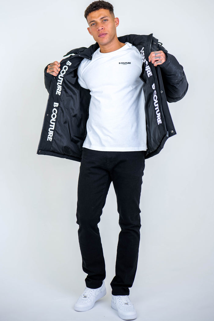 A man stands against a plain background, wearing the Arctic Puffer Jacket - Black with visible B COUTURE text on the inner lining. He pairs it with a white T-shirt, black jeans, and white sneakers, holding his jacket open to display the winter outerwear.