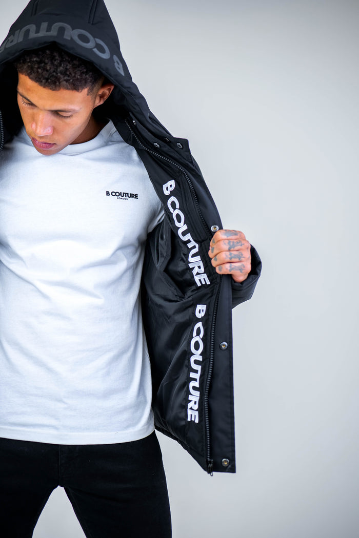 A man stands against a plain background, holding open his Arctic Puffer Jacket - Black to reveal the “B COUTURE” logo printed on the inner lining, while wearing a white B Couture t-shirt underneath.