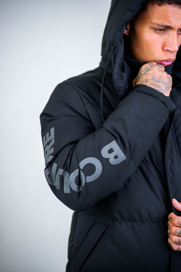 A person in the Arctic Puffer Jacket - Black, featuring bold sleeve lettering, holds the collar near their mouth. The winter outerwear photo focuses on their upper body and part of their face.
