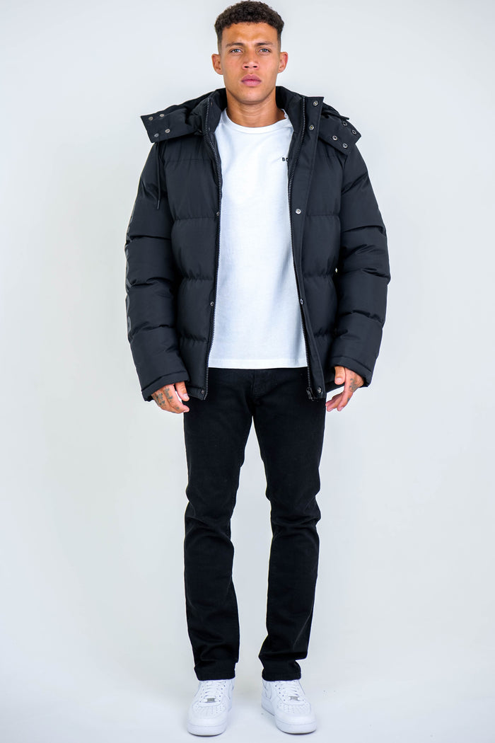 A young man stands facing forward in an Arctic Puffer Jacket - Black, paired with a white t-shirt, black pants, and white sneakers against a plain light gray background.