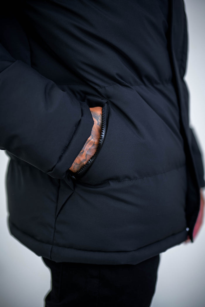 A close-up shows a person wearing the Arctic Puffer Jacket - Black, known for its thermal insulation, with their tattooed hand partially in the pocket. The plain background is out of focus.