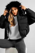 A woman with long blonde hair wears the Ashby Cropped Puffer Jacket - Black, featuring a fur-trimmed removable hood, over a light gray top and dark leggings, while sitting on a white block against a plain gray background.