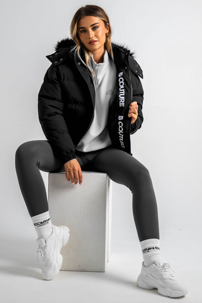 A woman sits on a white block wearing the Ashby Cropped Puffer Jacket - Black with faux fur trim and "COUTURE" text on the inside, paired with gray leggings, white sneakers, and white socks.