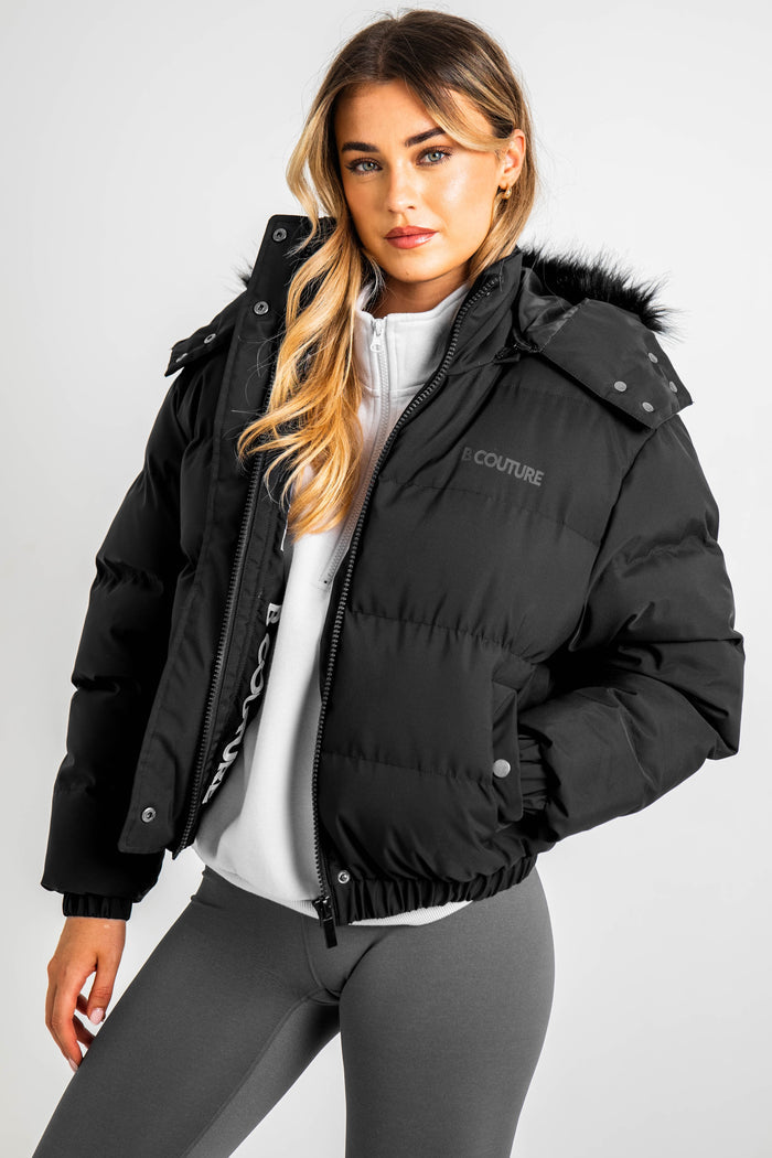 A woman models the Ashby Cropped Puffer Jacket in black, featuring synthetic insulation and a faux fur hood, layered over a white zip-up top and gray leggings. She poses against a plain background with one hand in her pocket, looking at the camera.