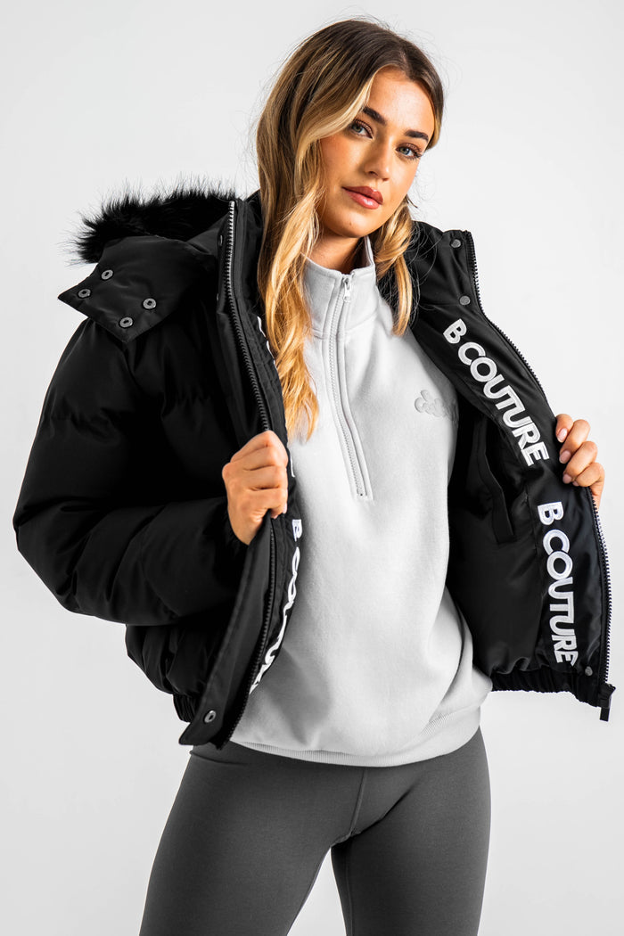 A woman with long, wavy hair wears the Ashby Cropped Puffer Jacket in black, featuring synthetic insulation and "B COUTURE" on the inside, over a light gray zip-up top and dark gray leggings against a plain light background.