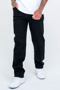A person wears Beckenham Straight Fit Stretch Denim Jeans in black, a white t-shirt, and white sneakers, standing with one hand in their pocket against a plain light background. Their upper face is not visible.