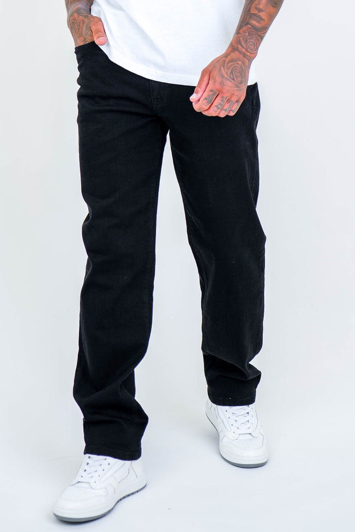 A person wears Beckenham Straight Fit Stretch Denim Jeans in black, a white t-shirt, and white sneakers, standing with one hand in their pocket against a plain light background. Their upper face is not visible.