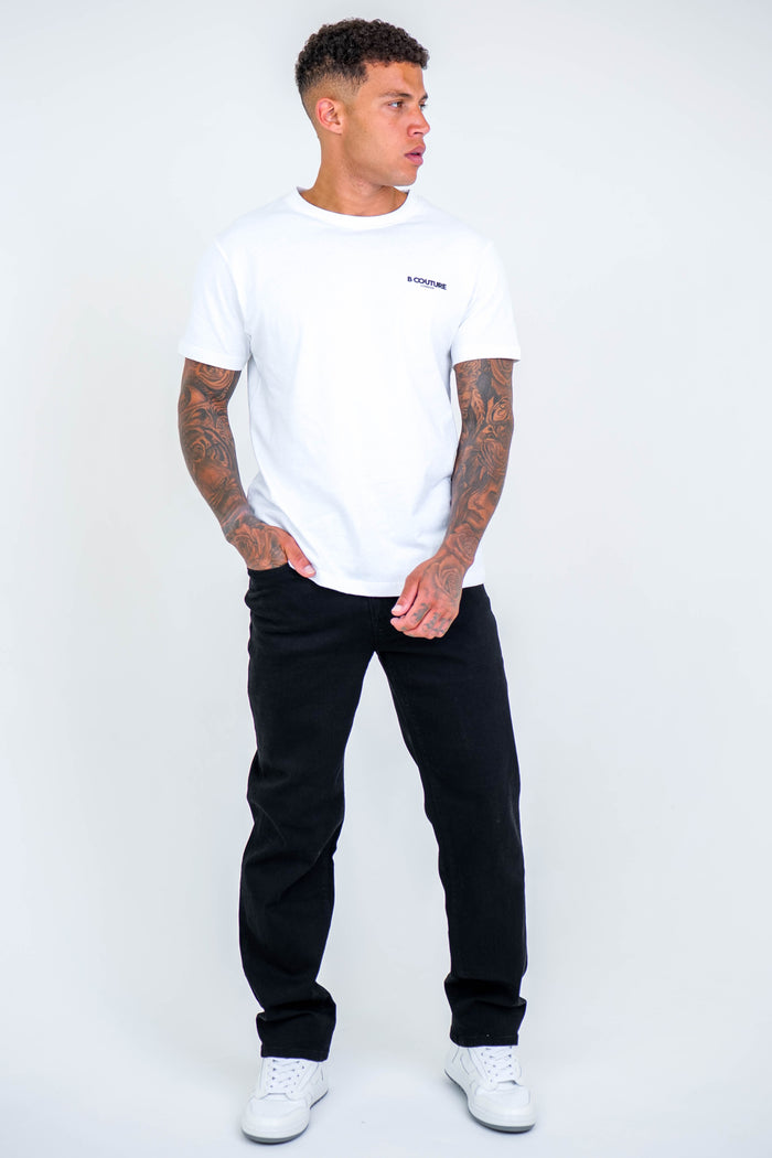 A man with tattoos wears a white t-shirt, Beckenham Straight Fit Stretch Denim Jeans in black, and white sneakers, standing against a plain white background while looking to his left.