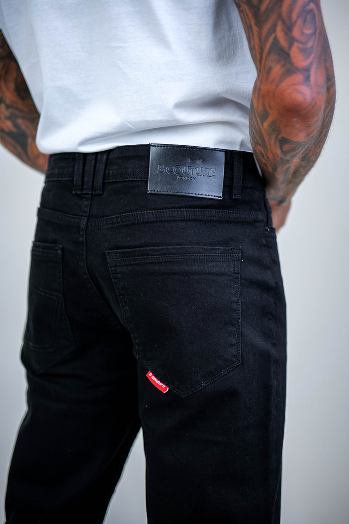 A person wearing a white t-shirt and Beckenham Straight Fit Stretch Denim Jeans - Black stands with their back to the camera, revealing tattooed arms and jeans with a black leather patch and small red tag on the back pocket.