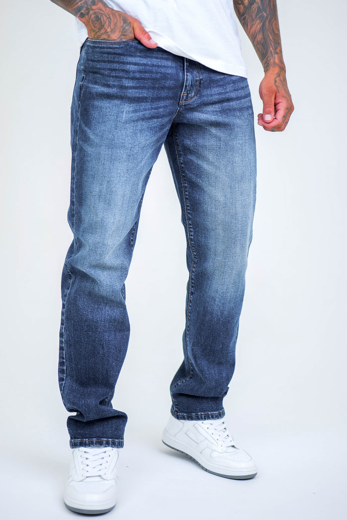 Wearing B Couture Beckenham Straight Fit Stretch Denim Jeans in Dark Blue with a white T-shirt and sneakers, a person stands with one hand in their pocket, showing only the lower half and highlighting the jeans' relaxed, comfortable fit.