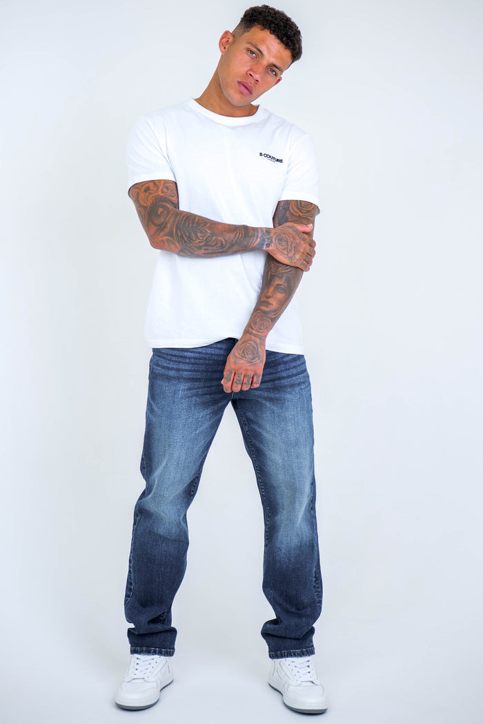 A man with tattoos wears a white t-shirt, B Couture Beckenham Straight Fit Stretch Denim Jeans in dark blue, and white sneakers. He stands against a plain light background with one arm crossed over his chest, looking at the camera.