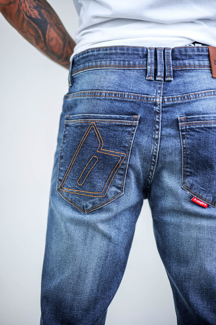 Close-up of a tattooed person wearing B Couture's Beckenham Straight Fit Stretch Denim Jeans in dark blue, featuring a bold “b” stitched on the back pocket and a red tag on the right pocket.