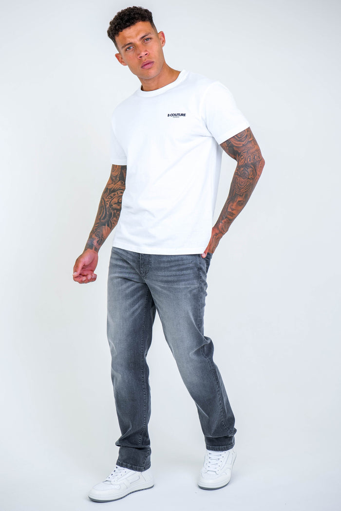 A man with short curly hair and arm tattoos stands against a white background, wearing a white t-shirt, B Couture Beckenham Straight Fit Stretch Denim Jeans in grey, and white sneakers. He looks slightly upward with a neutral expression.
