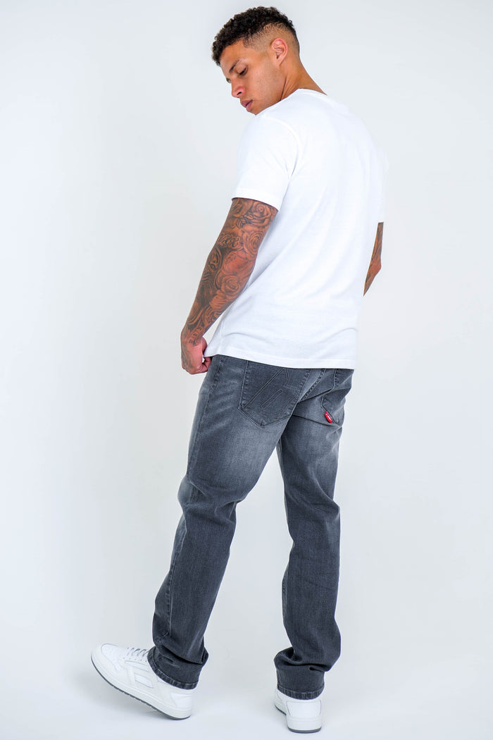 A tattooed man stands looking over his shoulder, wearing a white T-shirt, B Couture Beckenham Straight Fit Stretch Denim Jeans in Grey, and white sneakers against a plain white background.