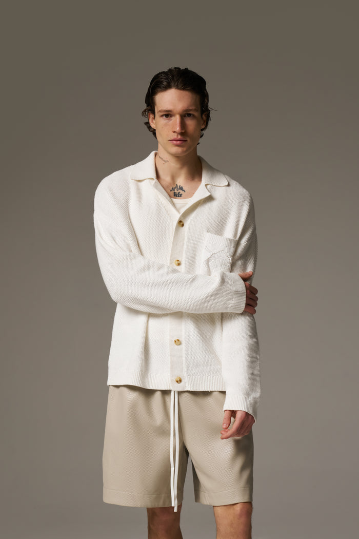 A man wearing a long sleeve crochet shirt and beige shorts against a neutral background.