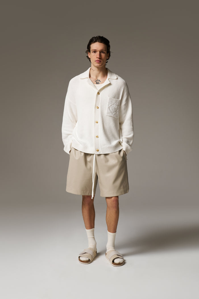 A man wearing a long sleeve crochet shirt and beige shorts against a neutral background.