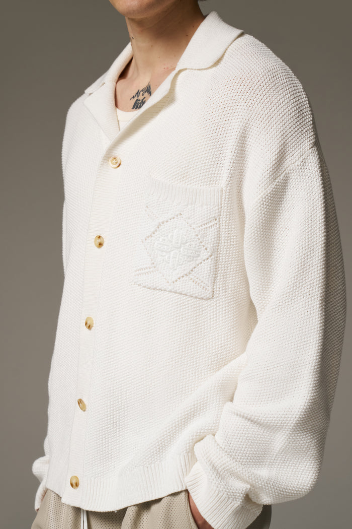 Close up detail short of man wearing a long sleeve crochet shirt and beige shorts against a neutral background.