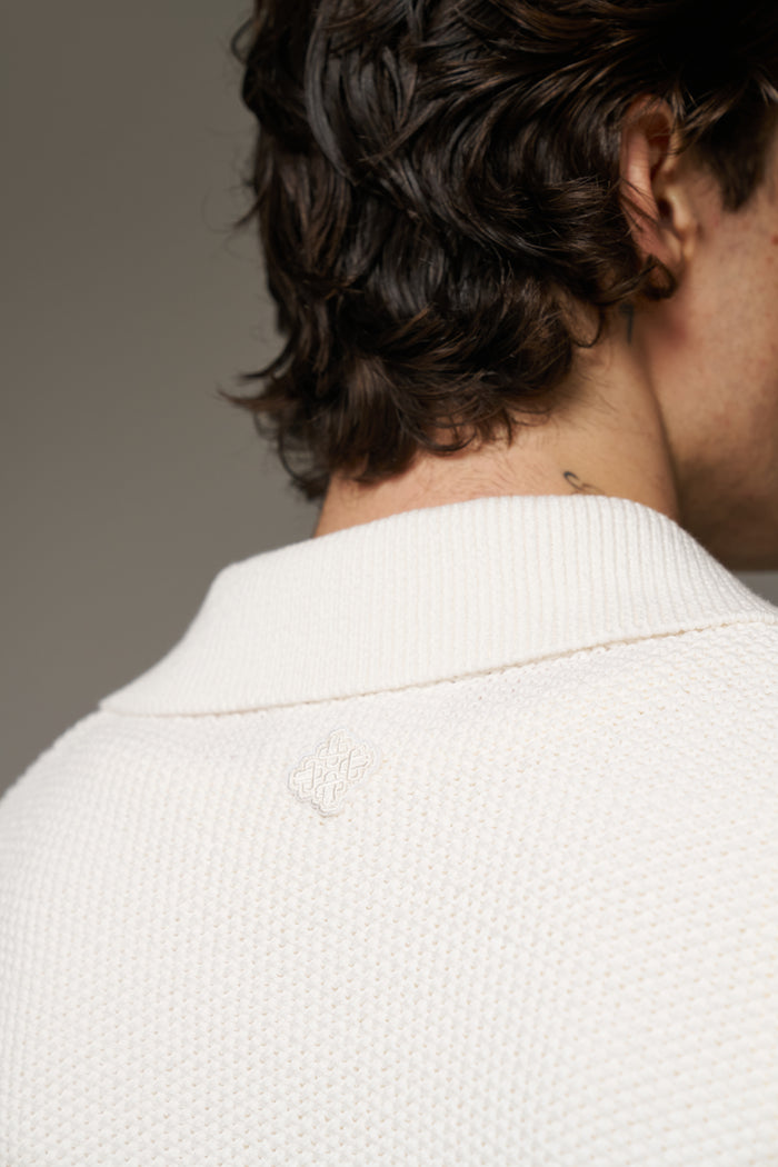 A close up detail shot of the back neckline of a man wearing an off white crochet shirt with an embroidered emblem.