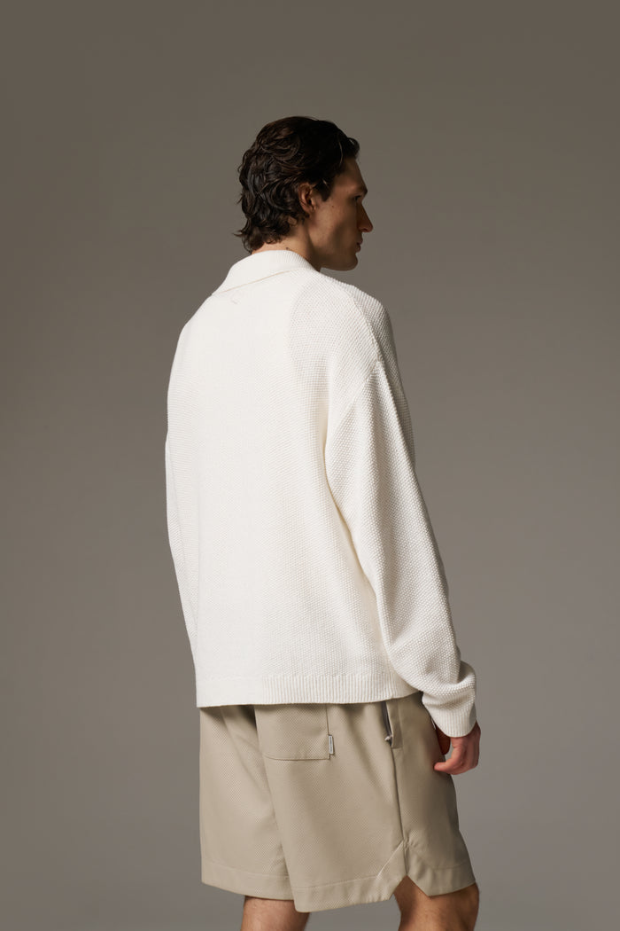 Back image of a man wearing an off white long sleeve crochet shirt and beige shorts against a neutral background.