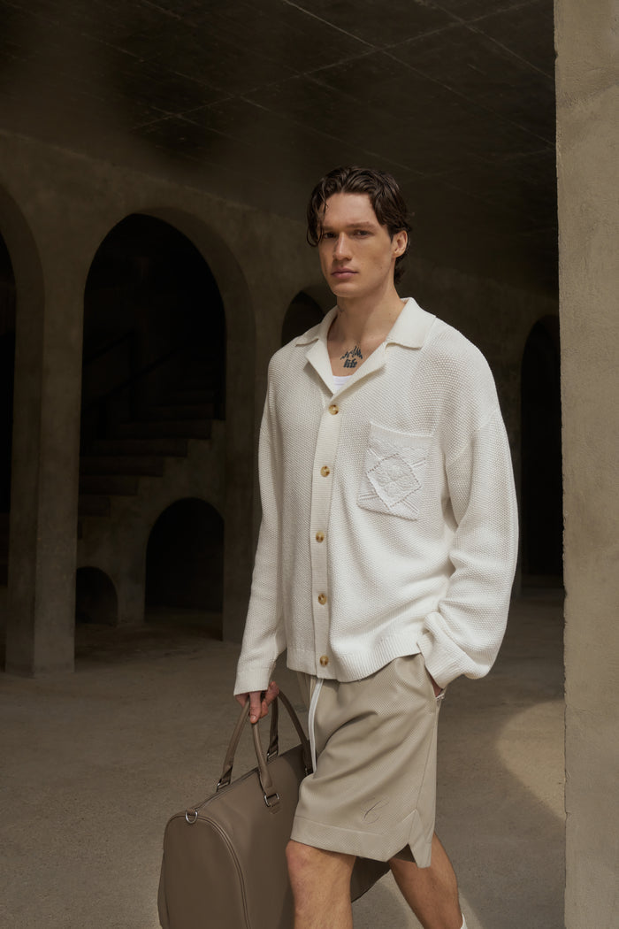A male model wearing an off-white long-sleeved shirt with a ribbed revere collar and a patch pocket on the chest, paired with matching shorts, holding a bag.