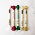 Five metallic DMC Light Effects embroidery floss skeins in festive holiday colors on a white marble background.