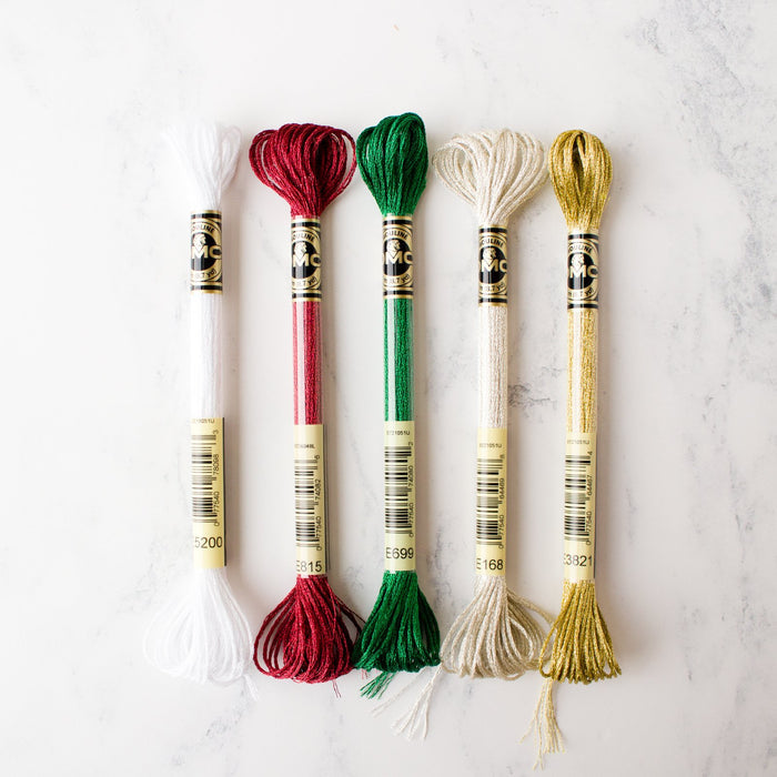 Five metallic DMC Light Effects embroidery floss skeins in festive holiday colors on a white marble background.