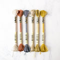 Six metallic DMC Light Effects embroidery floss skeins in neutral, copper, and gold shades on white marble.