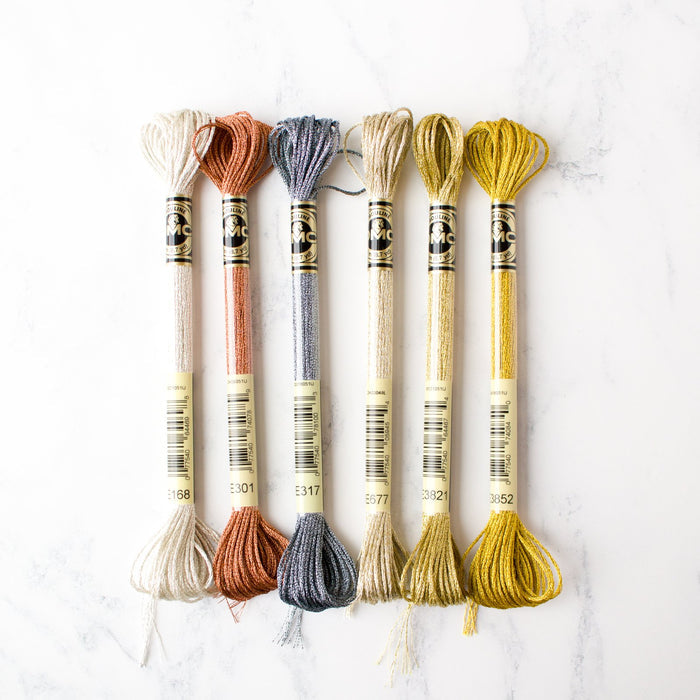 Six metallic DMC Light Effects embroidery floss skeins in neutral, copper, and gold shades on white marble.