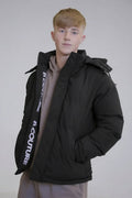 A young person with short blond hair models the Earlham Puffer Boys Jacket in black, featuring “COUTURE” text on the inner seam, layered over a taupe hoodie—an ideal winter style against a neutral background.