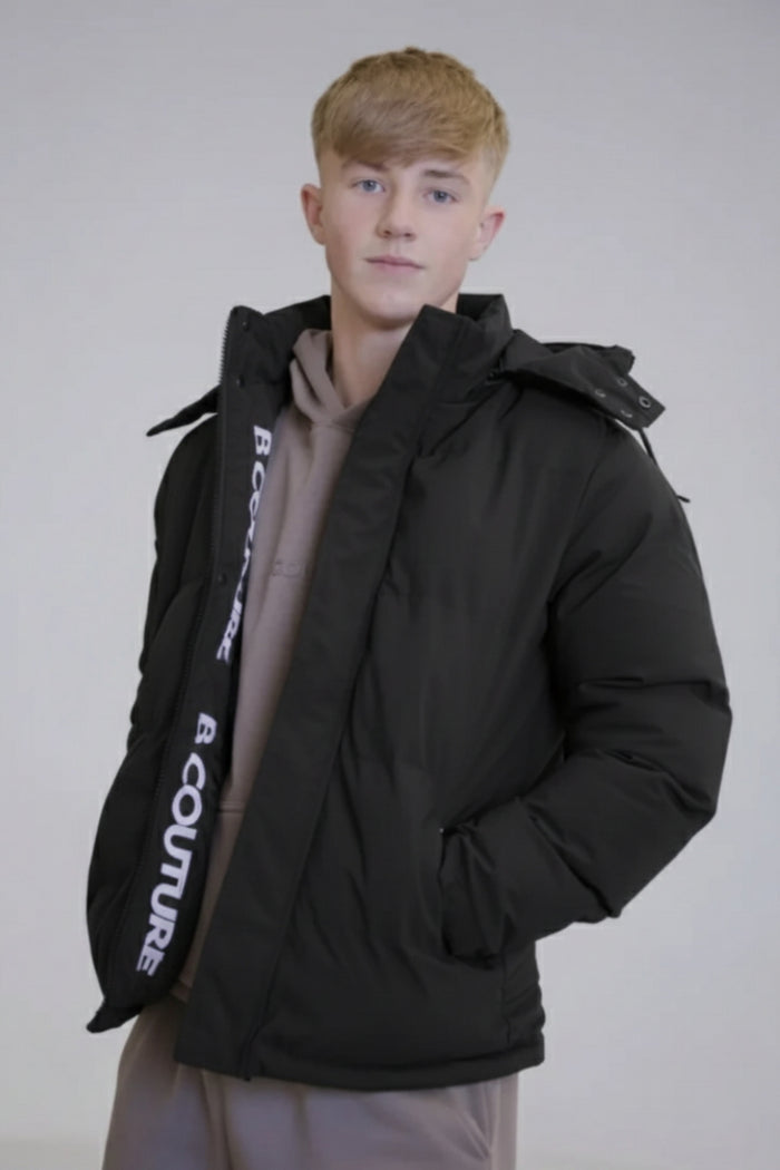 A young person with short blond hair models the Earlham Puffer Boys Jacket in black, featuring “COUTURE” text on the inner seam, layered over a taupe hoodie—an ideal winter style against a neutral background.