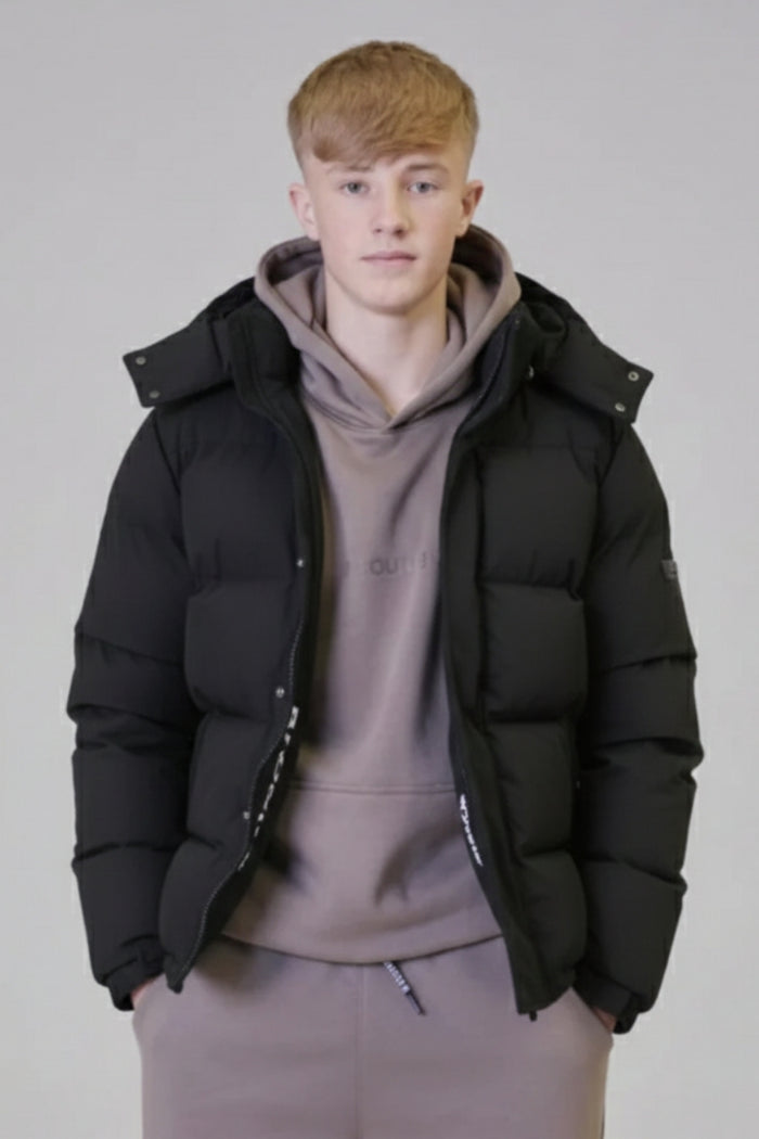 A young person with short blond hair wears the Earlham Puffer Boys Jacket in black over a taupe hoodie, standing against a plain light gray background and looking forward with a neutral expression.