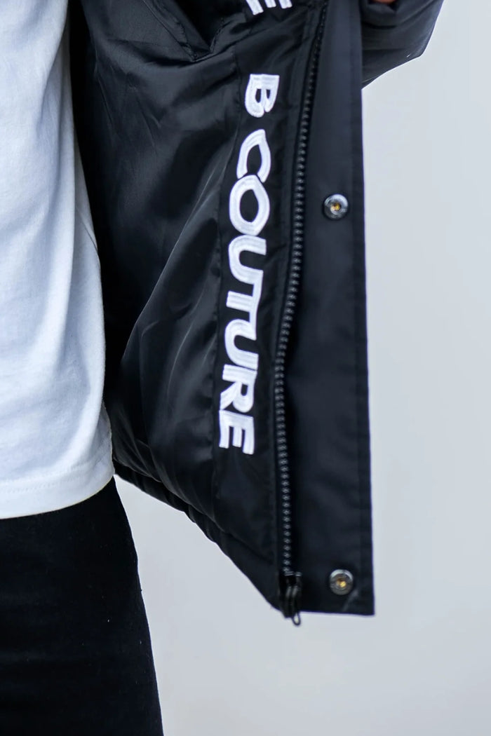 A close-up of a person wearing the Earlham Puffer Boys Jacket in black, partially unzipped to reveal bold white COUTURE lettering on the inner lining, styled with a white shirt and black pants for a sleek look.