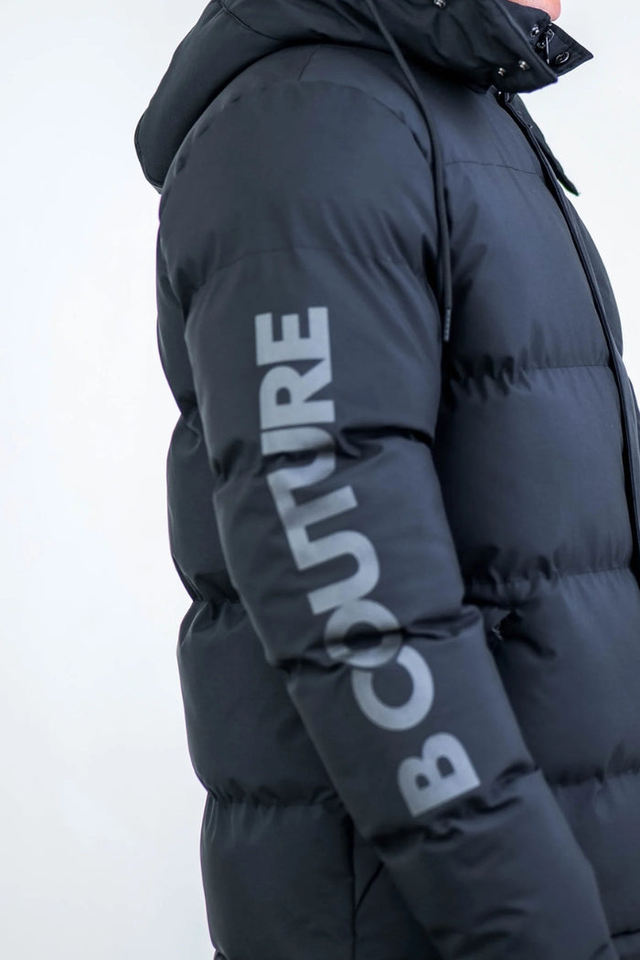 A person wears the Earlham Puffer Boys Jacket in black, featuring “COUTURE” in large gray letters on the sleeve, shown from an upper body side view against a plain background.