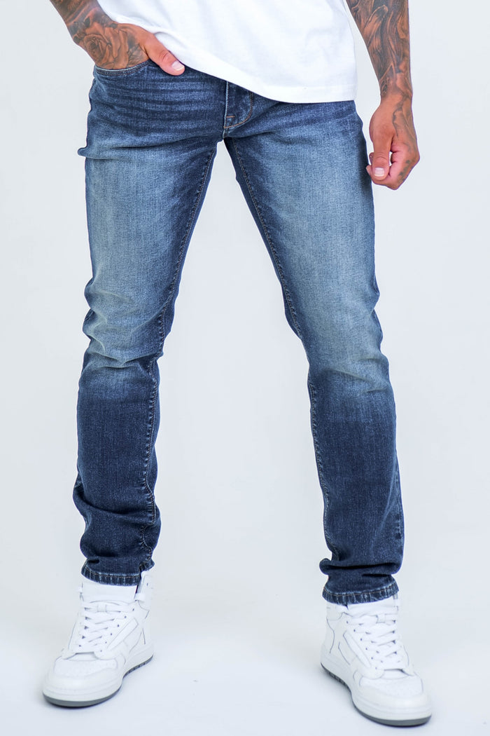 A person wearing Farnfield Taper Fit Stretch Denim Jeans in dark blue, a white T-shirt, and white sneakers stands with one hand in their pocket against a plain light background. Only the lower body is visible.