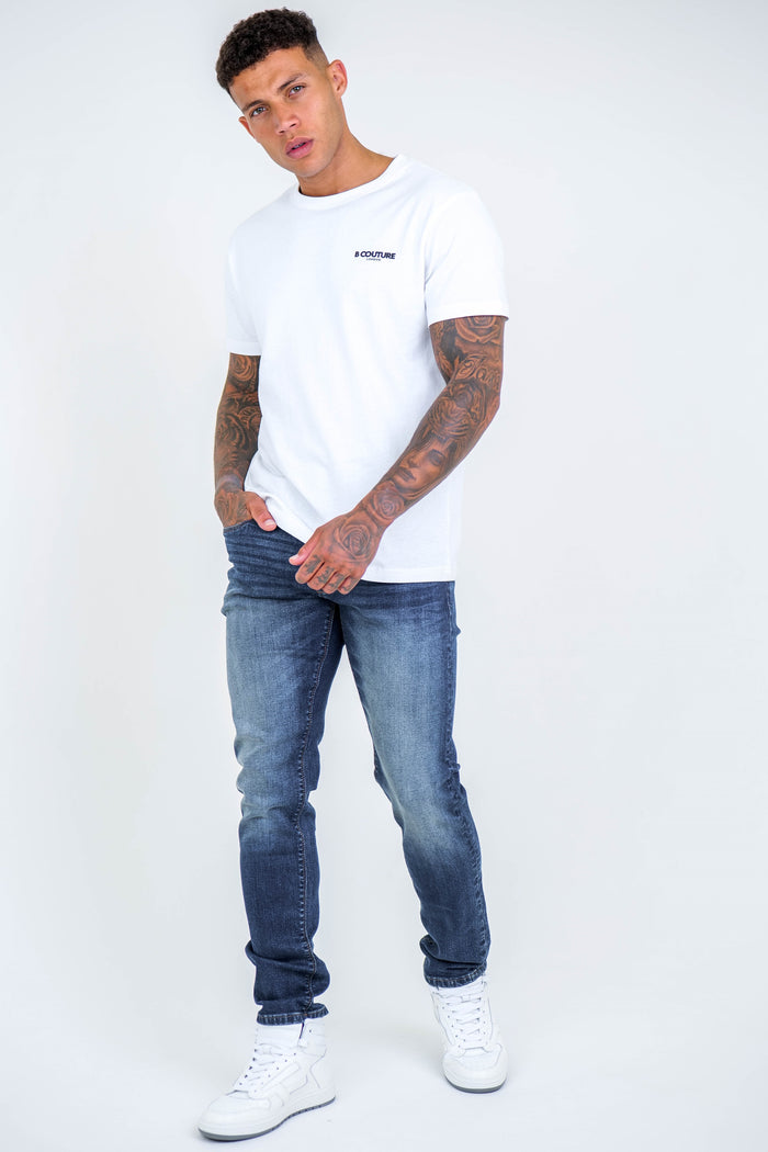 A tattooed man wearing a white T-shirt, Farnfield Taper Fit Stretch Denim Jeans in dark blue, and white sneakers stands against a plain light background, looking at the camera with a relaxed posture and one hand in his pocket.