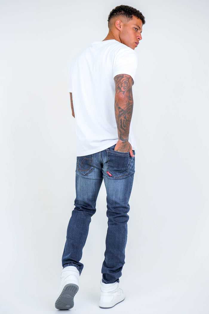 A man with tattoos wears a white T-shirt, white sneakers, and Farnfield Taper Fit Stretch Denim Jeans in dark blue. Facing away, he looks over his shoulder with his hands in his back pockets against a plain white background.