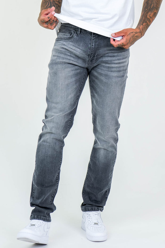 A person wearing a white t-shirt, Farnfield Taper Fit Stretch Denim Jeans - Grey, and white sneakers stands against a plain light background, holding the hem of their shirt with tattooed hands.