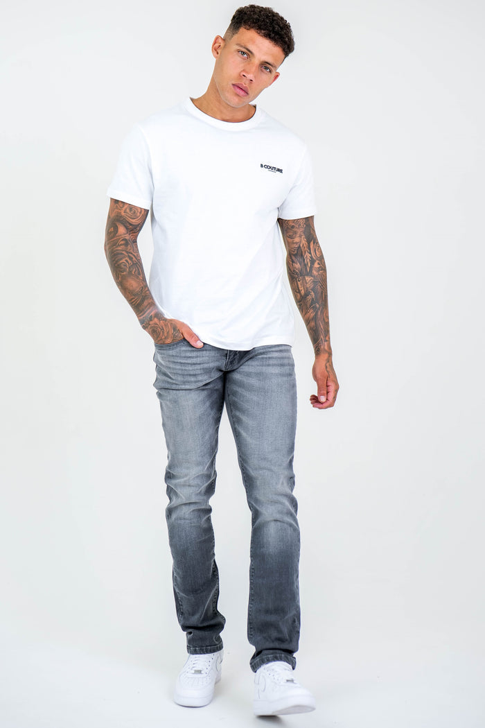 A man with short curly hair and full-sleeve tattoos wears a white t-shirt, Farnfield Taper Fit Stretch Denim Jeans in grey, and white sneakers, standing with one hand in his pocket against a plain white background.