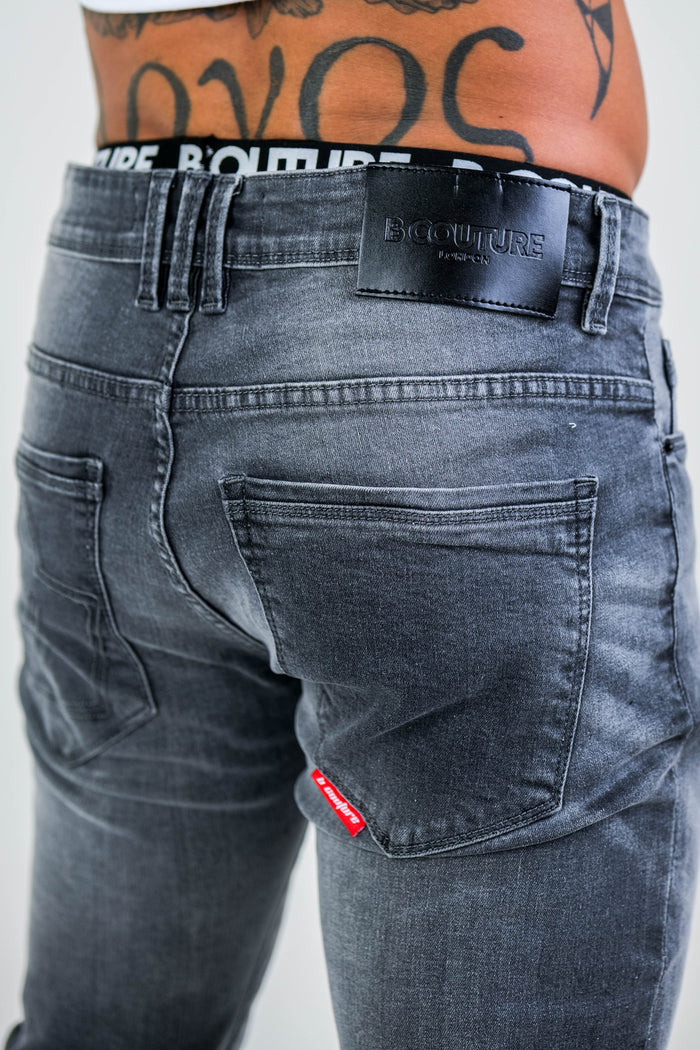 Someone wearing Farnfield Taper Fit Stretch Denim Jeans in grey, featuring a visible BCOUTURE PARIS label on the waistband. Part of a lower back tattoo peeks out above the slim fit stretch denim.