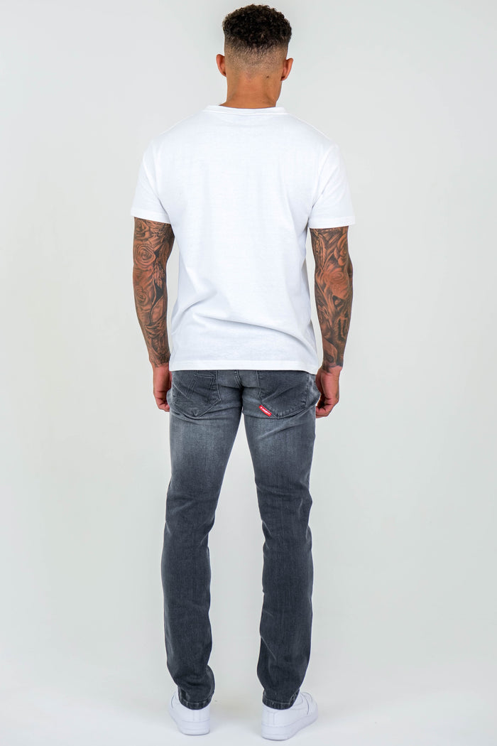 A man with short curly hair, visible arm tattoos, in a plain white t-shirt, Farnfield Taper Fit Stretch Denim Jeans - Grey, and white sneakers stands facing away from the camera against a plain light background.