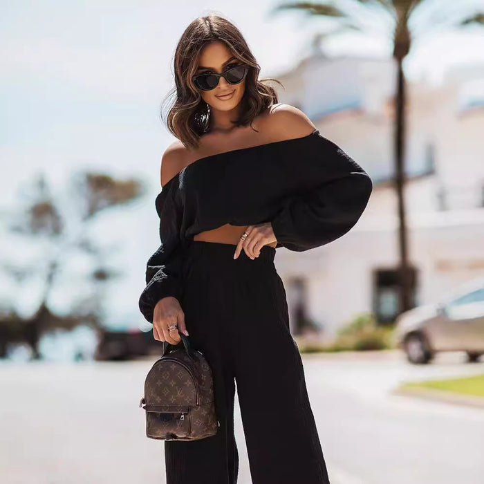 Woman in black off-shoulder top and pants holding a small backpack on a street.