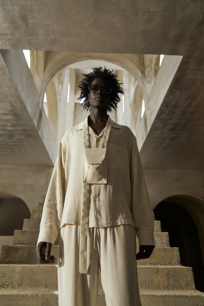 A male model wearing a beige textured woven shirt and matching pants, standing in a spacious indoor area with arched ceilings.