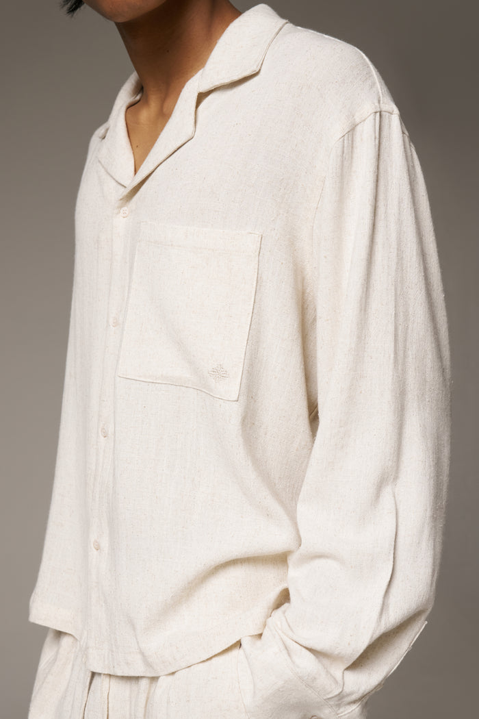 A close up shot of a man wearing a beige long sleeve shirt and matching pants against a neutral background.