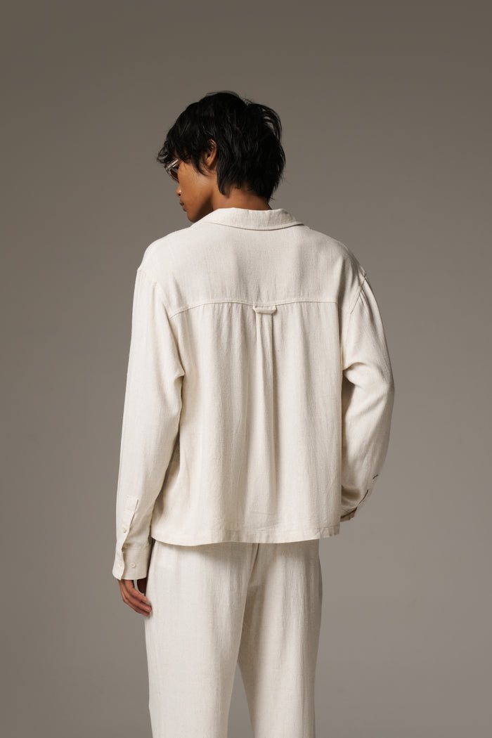 Back image of a man wearing a long sleeved linen shirt and patching pants, against a neutral background.