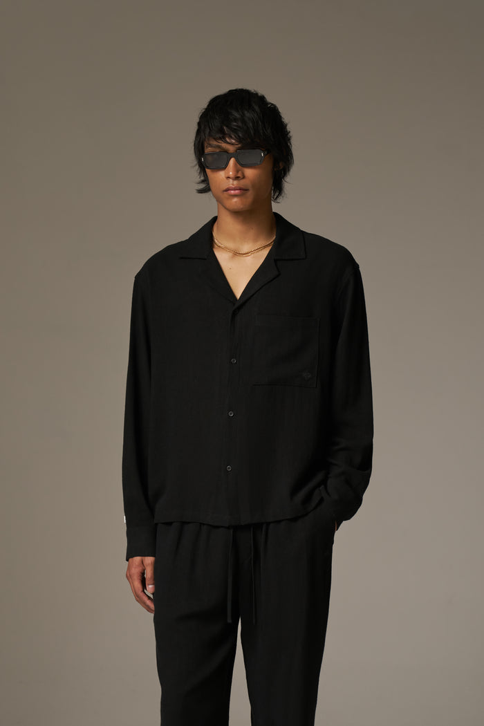 A man wearing a long sleeved shirt, with black sunglasses and pants against a neutral background.