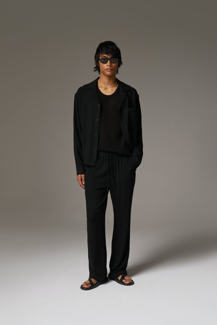 A man wearing a long sleeved shirt, with black sunglasses and pants against a neutral background.